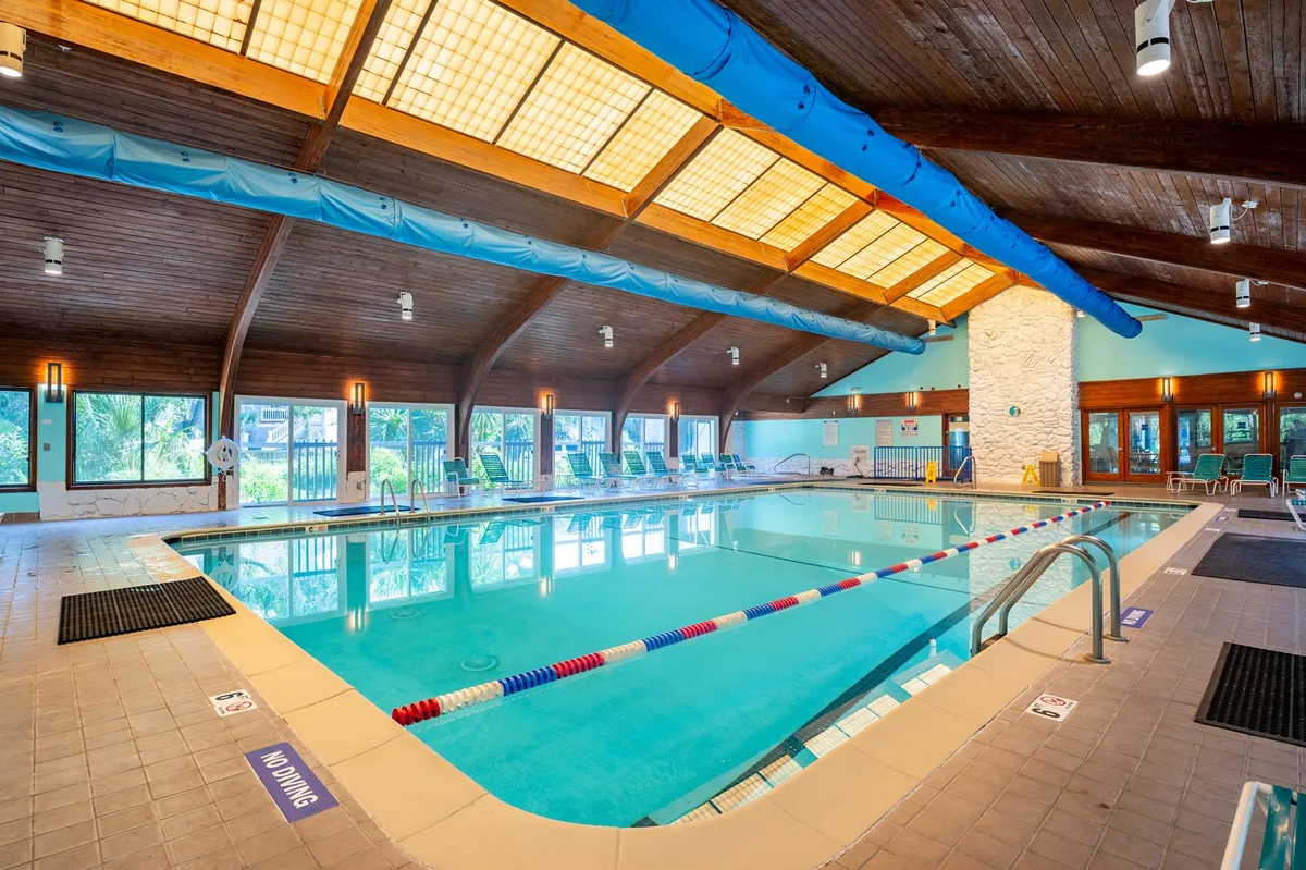 An inviting indoor swimming pool area with wooden ceilings, large windows that let in natural light, and poolside lounge chairs ideal for relaxing or socializing. The tranquil atmosphere highlights the year-round recreation available to guests.