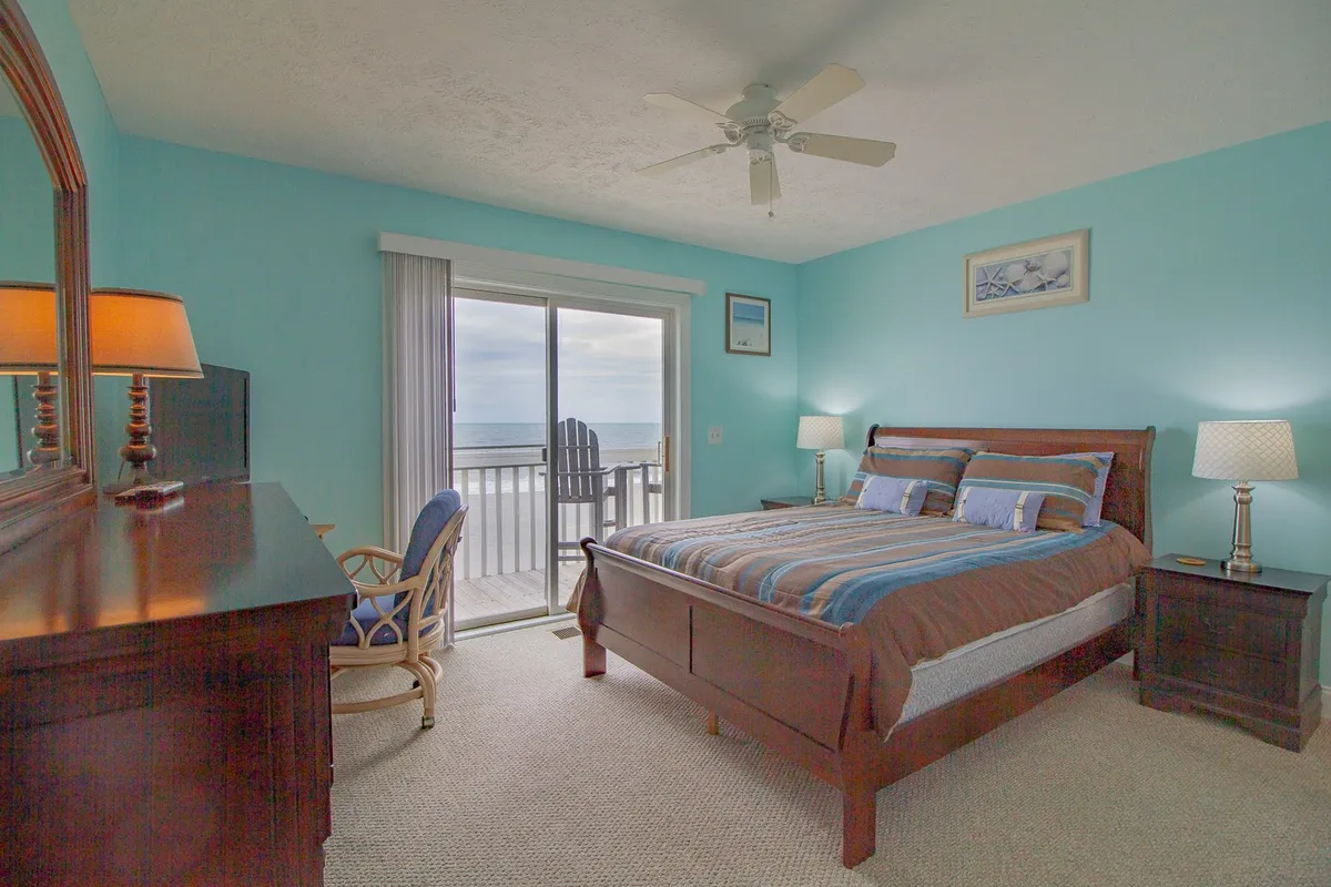 This image features a serene bedroom with a neatly made bed, a sliding glass door leading to a private deck or balcony, a dresser, and a lamp. The room feels calm and inviting, with natural light streaming in, suggesting a peaceful spot for rest or quiet work.