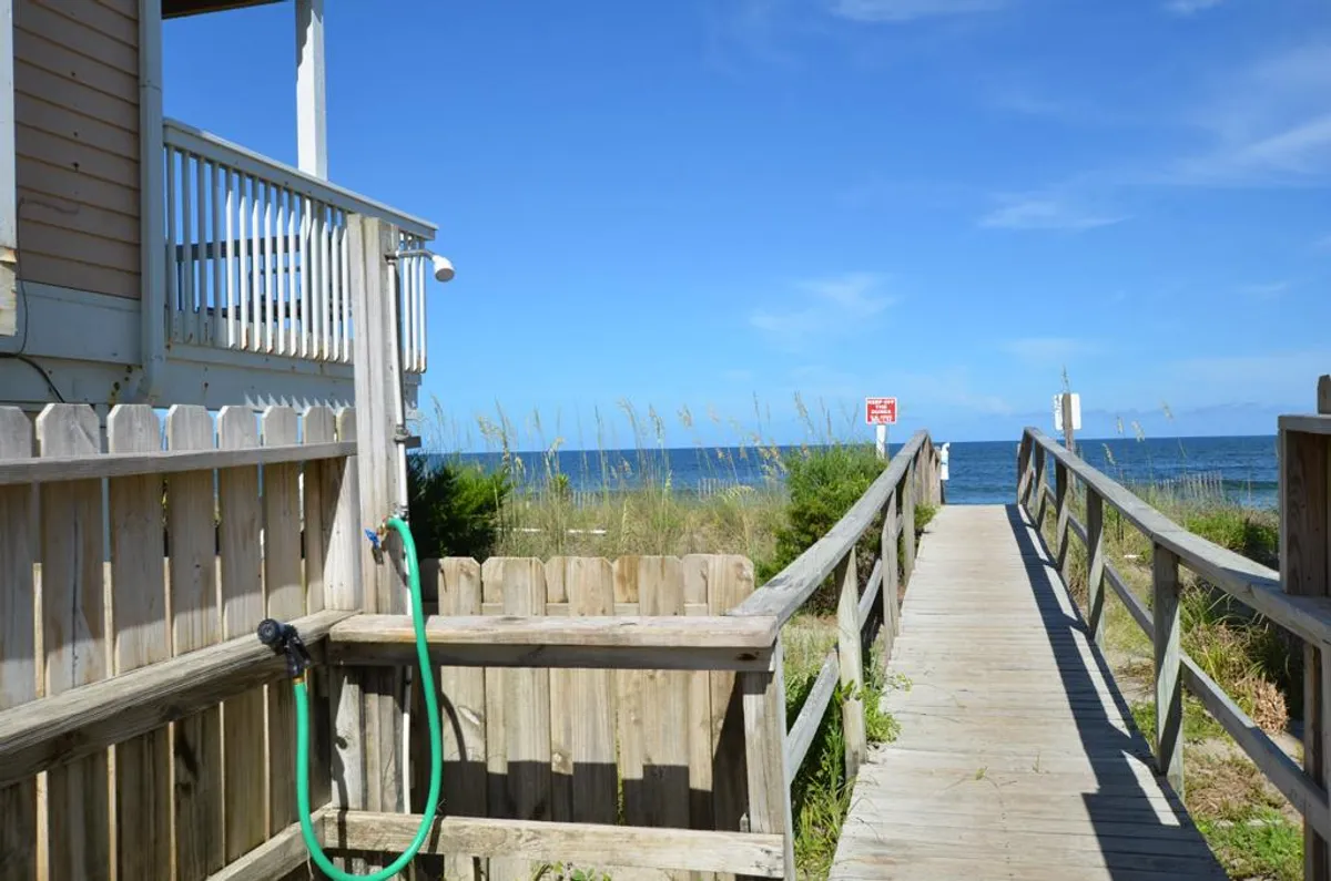 A scenic wooden walkway leading directly to the sandy beach, with lush greenery on either side and an inviting ocean view in the background. An outdoor shower is visible near the walkway, emphasizing convenience for families returning from the shore.