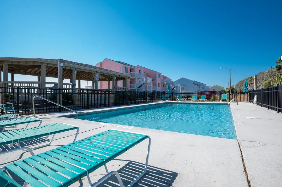 A bright exterior view of a sparkling swimming pool surrounded by comfortable lounge chairs, with nearby buildings framing the scene. The area is perfect for sunbathing, swimming, and socializing, embodying the lively, sunny vibe of a resort-style pool deck.