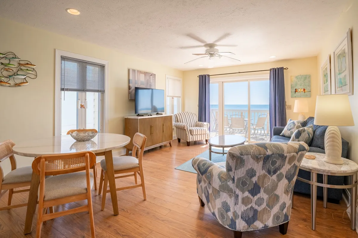 A tranquil, open-concept living and dining space with a dining table, cozy seating area, TV, and decorative wall art. Large windows offer a clear ocean view, seamlessly blending indoor comfort with the spectacular seaside scenery.