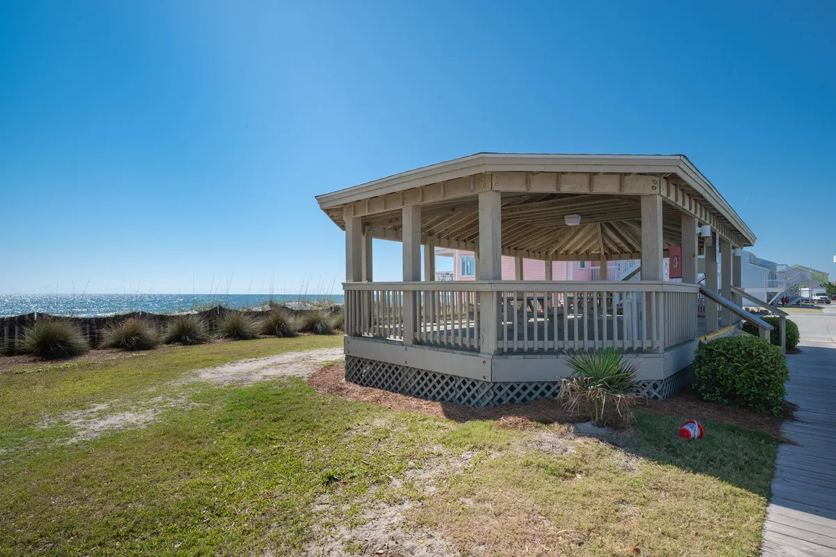 A tranquil gazebo overlooks the ocean, surrounded by lush greenery. The structure is set slightly above the sand and provides a sheltered spot for relaxing or socializing while taking in panoramic coastal views.