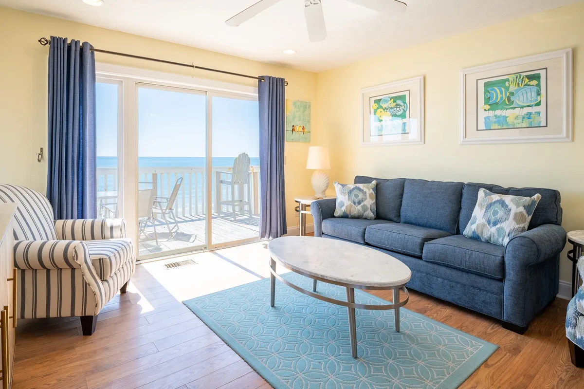 A bright and cheerful living room featuring a plush sofa and coffee table set in front of a large window, with blue curtains framing an expansive ocean view. The space is bathed in natural sunlight, emphasizing the connection to the outdoors.