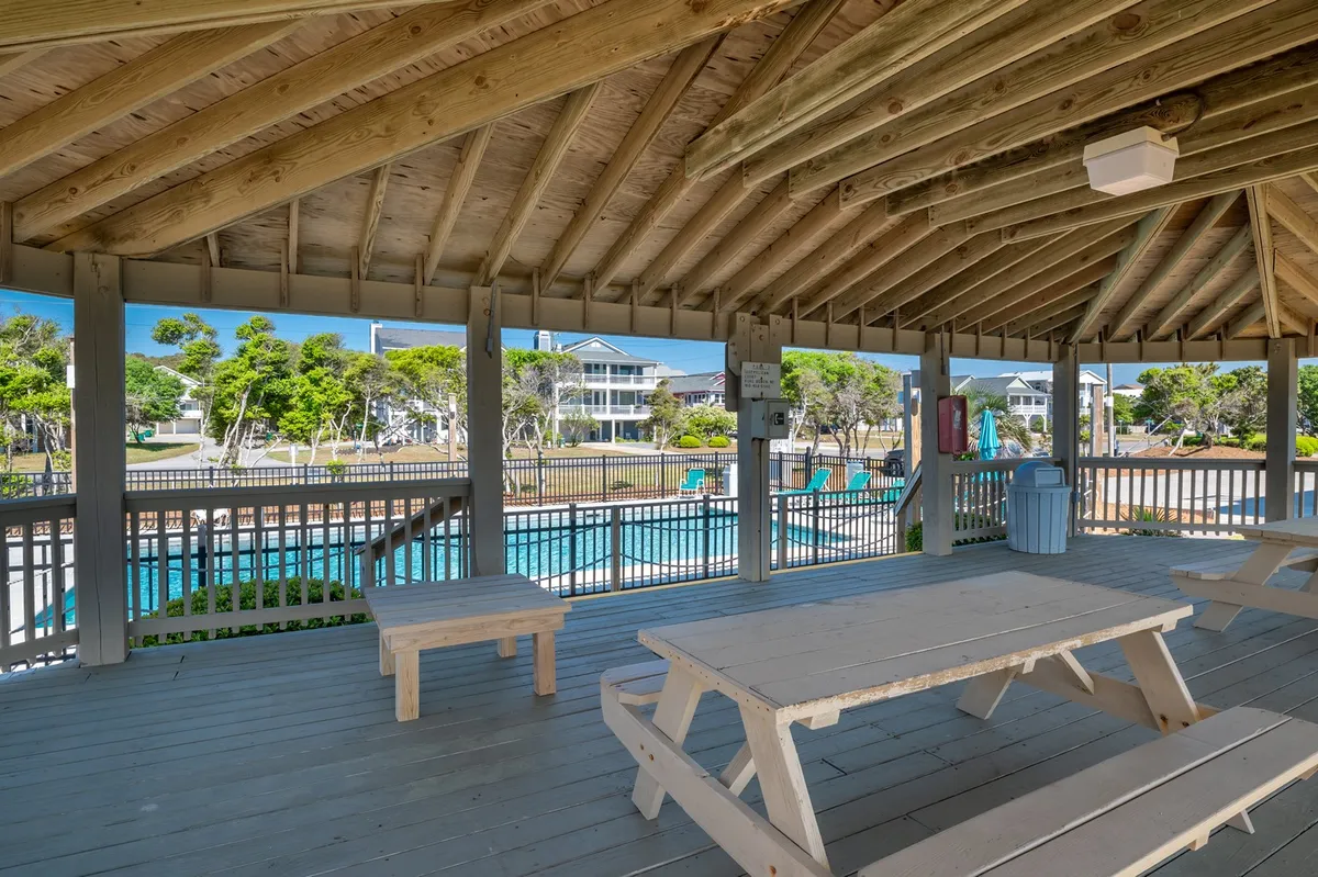 A vibrant outdoor scene featuring picnic tables beside a sparkling swimming pool, accented by wooden beams and lush landscaping. The area is set up for social gatherings, outdoor dining, and poolside relaxation, creating an atmosphere of cheerful community.
