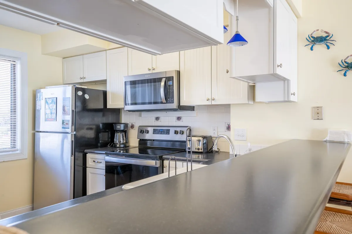 The image focuses on a cozy, functional kitchen scene with a black refrigerator, stainless steel microwave, white cabinetry, a standout blue pendant light, and playful decorative crabs on the wall. The space is clearly designed for both cooking and social interaction, with plenty of counter space and ambient natural light.