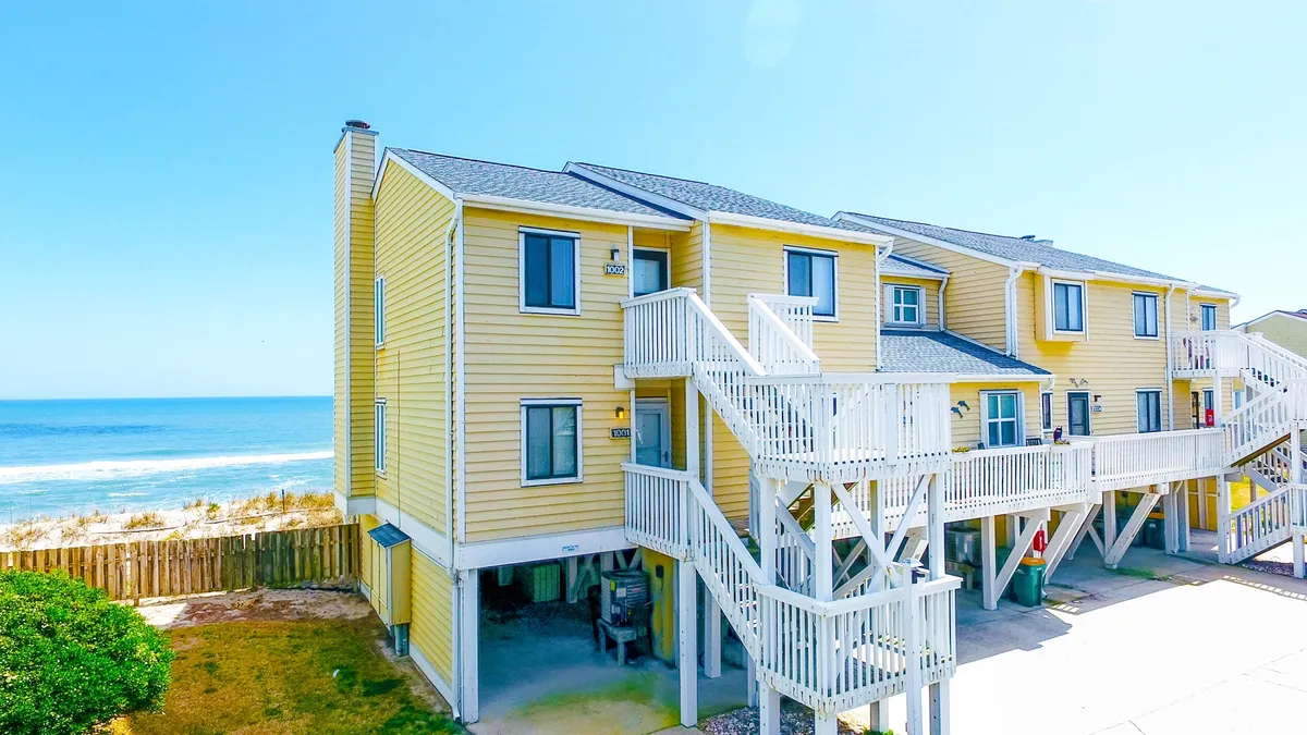 A cheerful exterior view showing the yellow facade of the building, a staircase leading to the upper levels, and a backdrop of the ocean. The porch offers space for relaxing while the proximity to the shoreline is clearly visible, evoking the coastal lifestyle and romance of being steps from the beach.