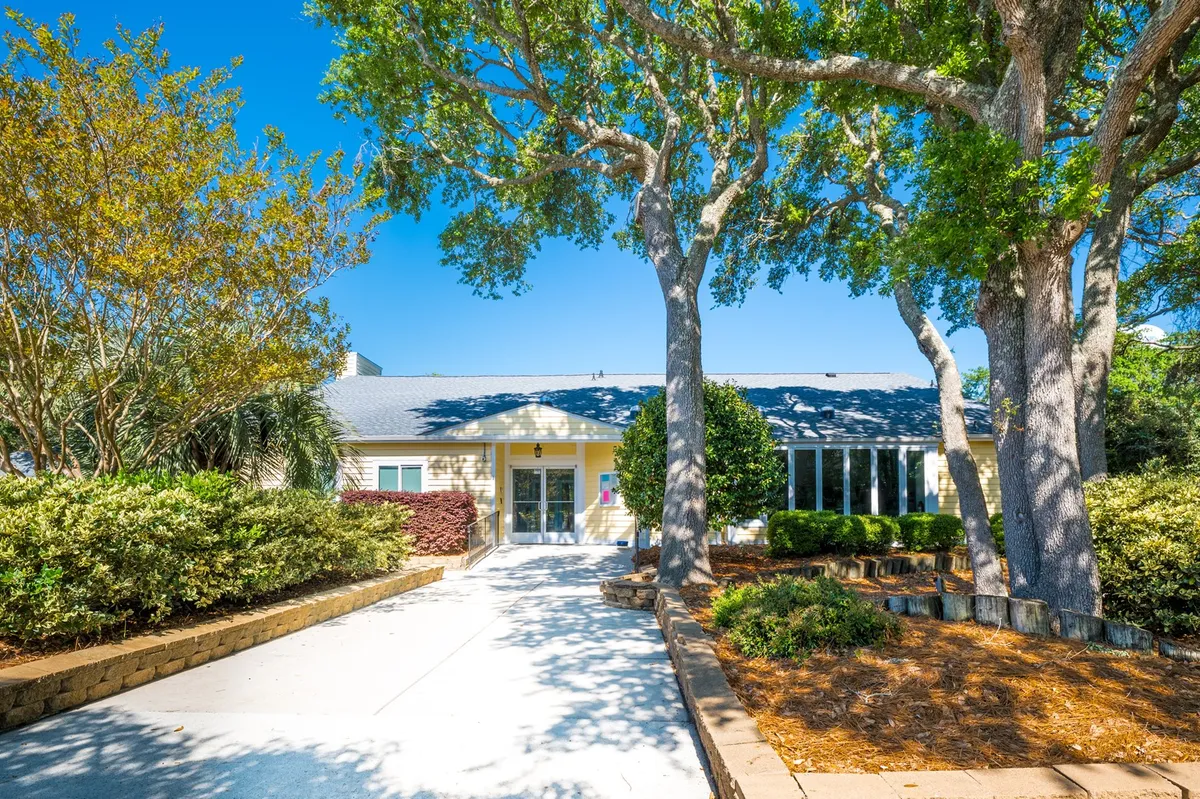A welcoming entrance pathway winds through large trees and manicured shrubs, leading to the building facade. The dappled shade creates an inviting setting for outdoor gatherings or peaceful moments amid nature.