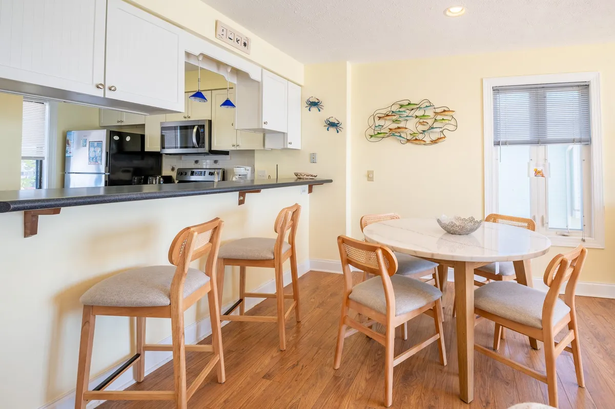 A bright and inviting interior highlighting a bar counter adjacent to a circular dining table, adorned with decorative wall art. The bar counter visually connects the kitchen to the living and dining areas, suggesting a social hub perfect for casual bites, drinks, and entertaining guests.