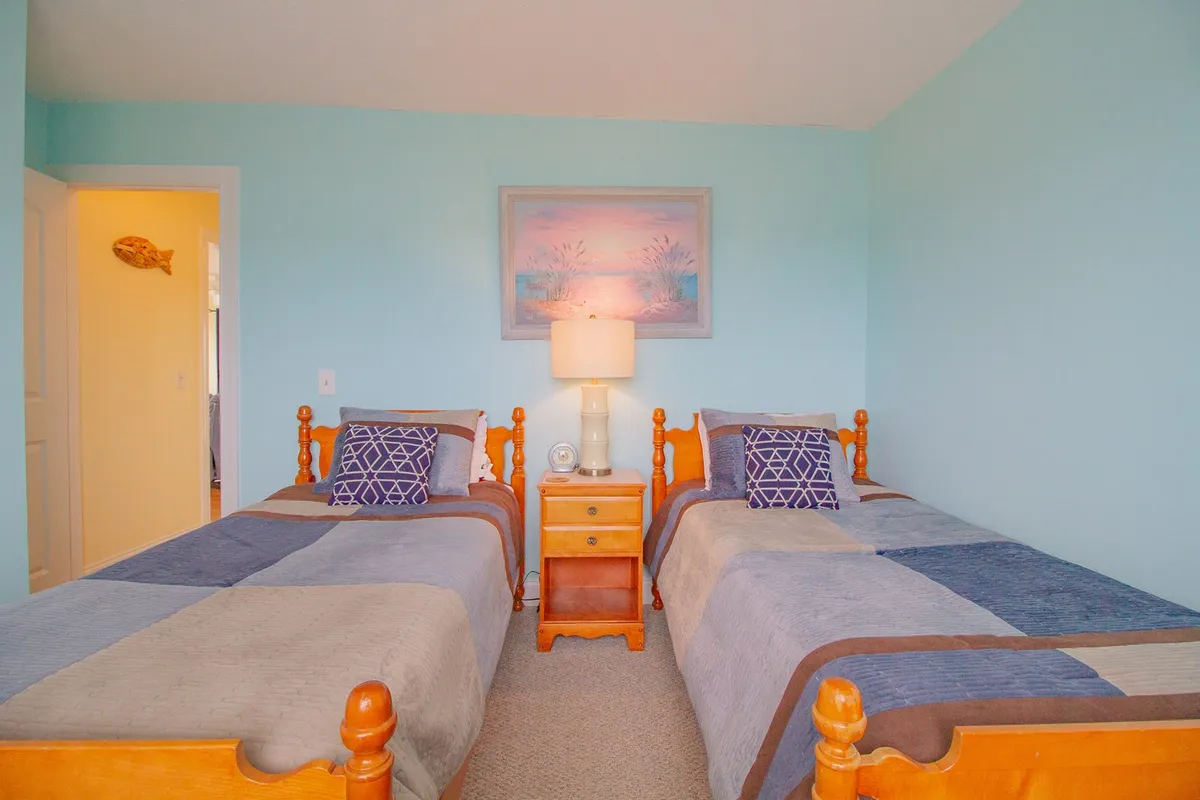 This image shows a second bedroom with two twin beds in wooden frames, a nightstand with a lamp, and decorative wall art. The space is cozy and organized, making it suitable for children or friends to unwind after a day of activities.