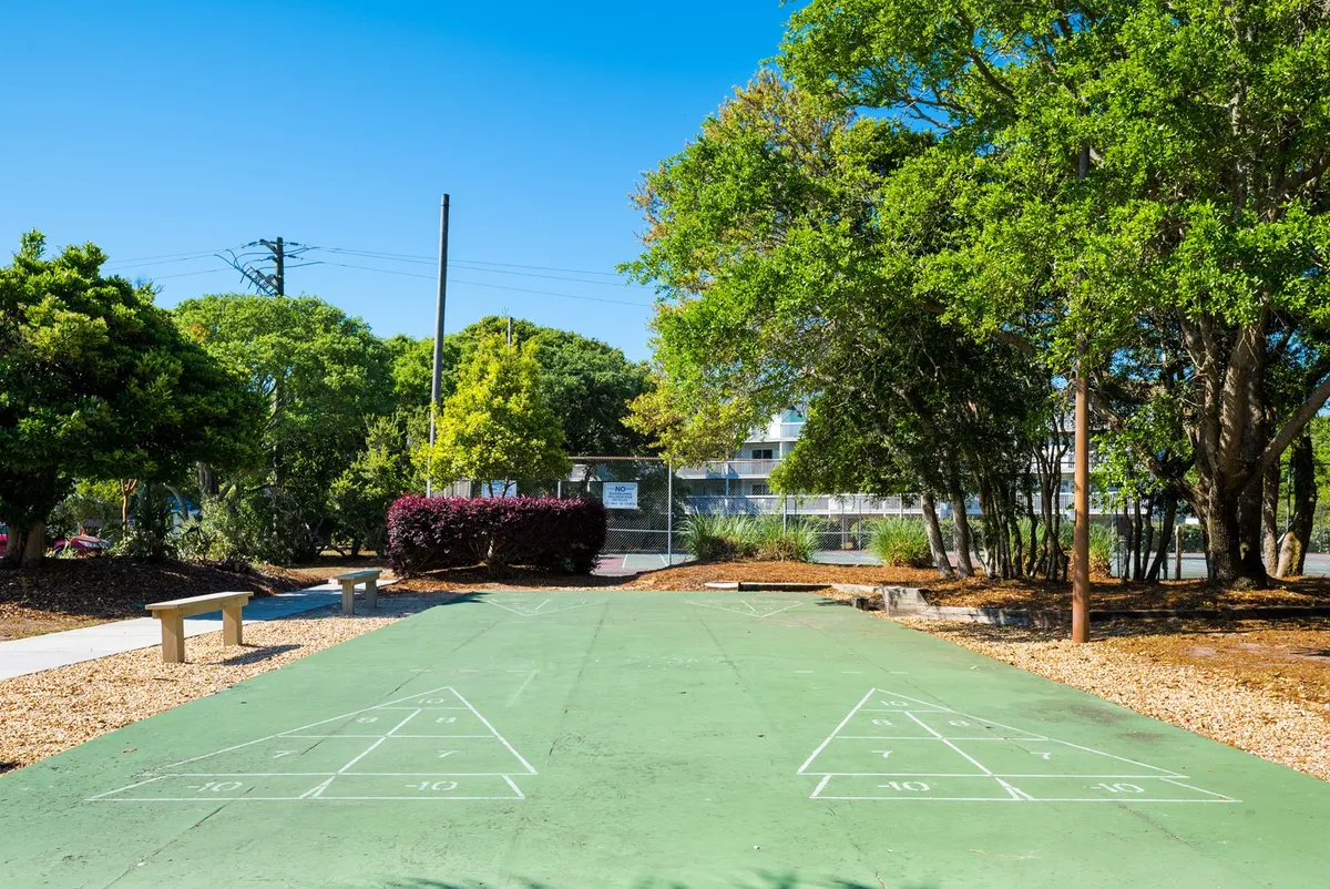 An inviting shuffleboard court sits among mature trees and benches, offering a relaxed, social space for guests to play and unwind outdoors. The scene evokes friendly competition and community engagement.